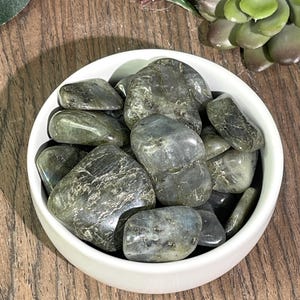 May include: A white bowl filled with tumbled labradorite stones. The stones are a dark gray color with iridescent flashes of blue and green.