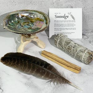 May include: An iridescent abalone shell resting on a wooden tripod stand, a bundle of dried white sage tied with string, a single brown and black patterned feather, a wooden stick, and an instructional card titled How to Smudge.