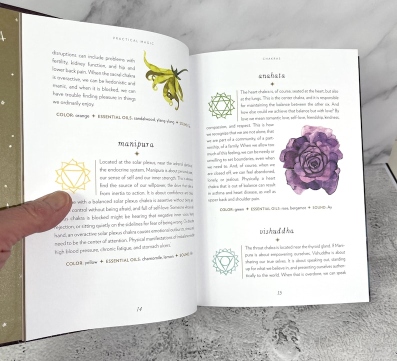 May include: A book open to a page about the Anahata chakra, the heart chakra. The page features a green watercolour illustration of a rose with a green and white mandala symbol. The text on the page reads "Anahata" and "The heart chakra is, of course, seated at the heart, but also at the lungs. This is the centre chakra, and is responsible for maintaining the balance between the other six. And how else could we achieve that balance but with love? By love we mean romantic love, self-love, friendship, kindness, compassion, and respect."