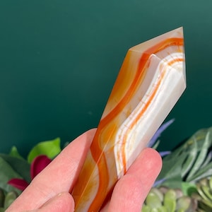 May include: A polished, orange and white banded agate crystal point. The crystal has a faceted top and is held in a hand against a green background with leafy plants. The agate has a translucent quality, with the orange bands creating a striking visual pattern.