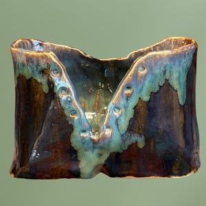 May include: A ceramic vase with a unique V-shaped design. The vase is glazed in shades of brown, green, and blue, with a textured surface.