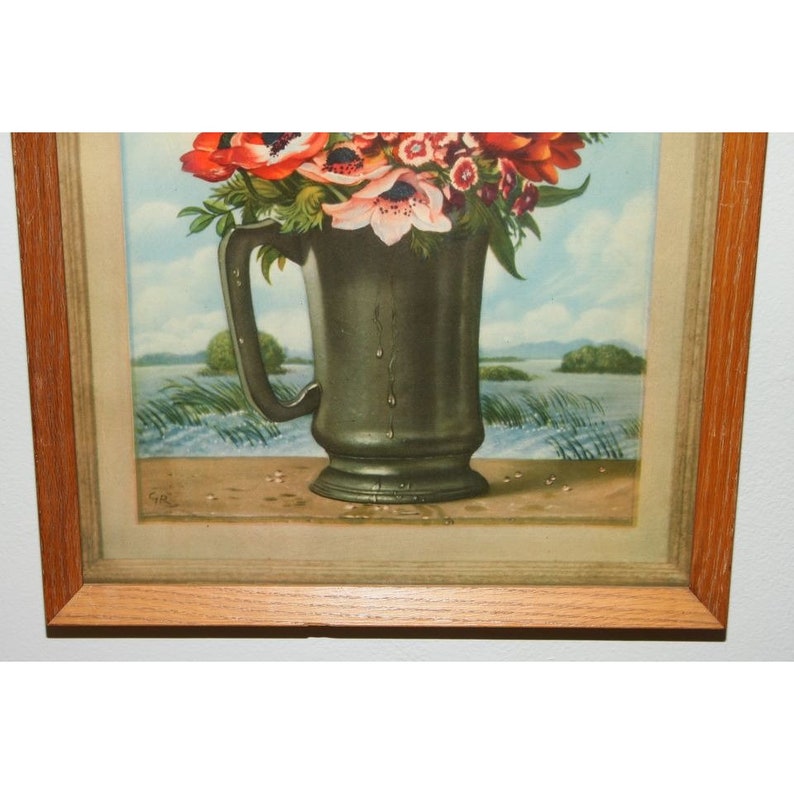 May include: A framed print of a still life featuring a pewter pitcher filled with red, pink, and white flowers. The pitcher is sitting on a table with a light brown surface. The background is a watercolor painting of a lake with green trees and a blue sky.