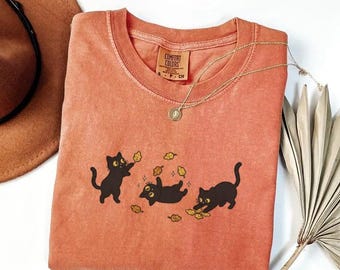 May include: Coral-colored Comfort Colors t-shirt featuring a playful graphic of three black cats with yellow eyes and autumn leaves. A gold necklace with a pendant rests on the shirt. The shirt is folded, with a brown hat visible in the background.