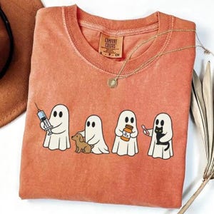May include: A coral-colored Comfort Colors t-shirt featuring four cartoon ghosts. One ghost holds a syringe, another a small dog, a third a pill bottle, and the last a black cat. A gold necklace with a pendant is draped across the shirt.