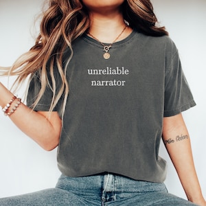 May include: A dark gray t-shirt with the text "unreliable narrator" printed in white on the front. The shirt is being worn by a person with long blonde hair and a gold necklace.