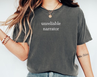 May include: A dark gray t-shirt with the text "unreliable narrator" printed in white on the front. The shirt is being worn by a person with long blonde hair and a gold necklace.