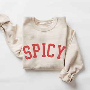 May include: A cream-colored sweatshirt with the word "SPICY" printed in red block letters across the front. The long sleeves are rolled up. The sweatshirt is laid flat on a white surface.