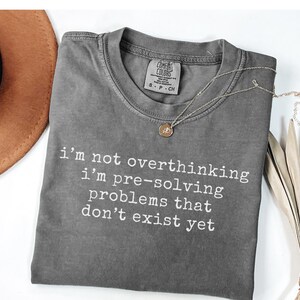 May include: A charcoal gray short-sleeve t-shirt with a crew neckline and the white printed text: i'm not overthinking i'm pre-solving problems that don't exist yet. The shirt features a Comfort Colors brand label at the inner neck area.