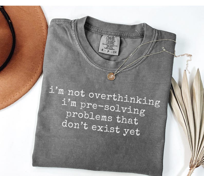 May include: A charcoal gray short-sleeve t-shirt with a crew neckline and the white printed text: i'm not overthinking i'm pre-solving problems that don't exist yet. The shirt features a Comfort Colors brand label at the inner neck area.