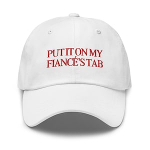 May include: White baseball cap with the embroidered text "PUT IT ON MY FIANCÉ'S TAB" in red. The cap has a curved brim and a button on top. A casual accessory.