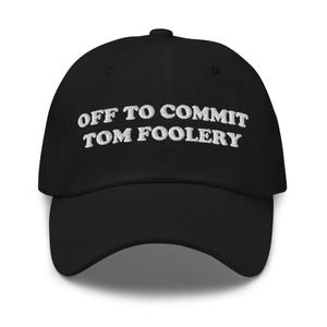 May include: Black baseball cap with white embroidered text that reads "OFF TO COMMIT TOM FOOLERY". The cap has a curved brim and a fabric strap closure on the back. A casual accessory.