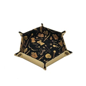 May include: A hexagonal dice tray with a tan exterior and a black interior patterned with gold-colored fantasy-themed illustrations. The tray has snap closures at each corner.
