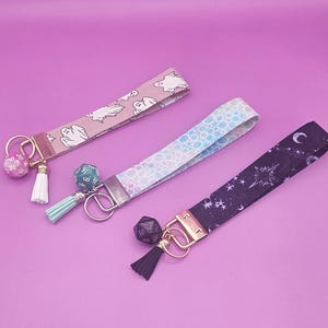 May include: Three keychains with different fabric patterns and charms. The first keychain has a pink and white ghost pattern, the second has a light blue and white pattern, and the third has a black and white star pattern. Each keychain has a gold key ring and a charm with a dice.