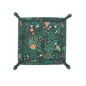 May include: A square, dark green fabric tray with a whimsical forest pattern. The design includes mushrooms, flowers, and crescent moons in shades of orange, white, and green. The tray has folded corners for easy storage and use.