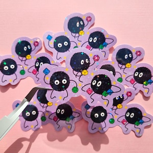 May include: A collection of stickers featuring a black, fluffy character with white eyes and a smiling expression. Each sticker has a light purple outline and is decorated with colorful geometric shapes. A pair of tweezers is in the foreground.