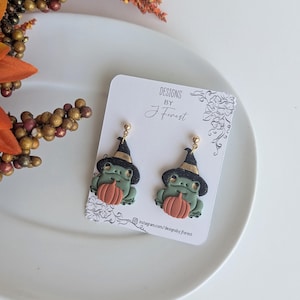 May include: A pair of Halloween-themed earrings featuring green frogs wearing black witch hats and holding orange pumpkins. The earrings are on a white card with the text "DESIGNS BY J. Forest" and a floral design.