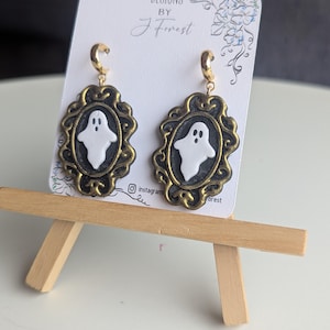 May include: A pair of dangle earrings featuring a white ghost figure centred within an oval black and gold ornate frame. The earrings hang from gold-toned hoops and are displayed on a white card labelled Designs by J Forest with floral art.