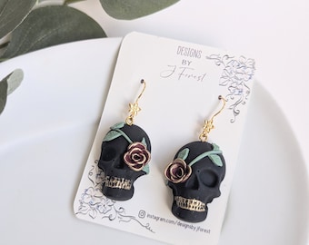Puede incluir: Pendientes de calavera negros con detalles dorados y una rosa roja. Los pendientes están en una tarjeta blanca con el texto "DESIGNS BY J Forest" y un identificador de redes sociales. Los pendientes cuelgan de ganchos dorados.
