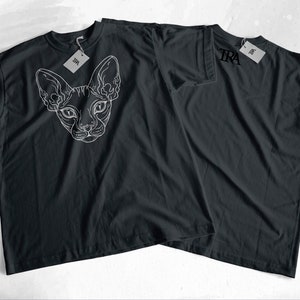 May include: Two black t-shirts with white graphic designs. One shirt features a line drawing of a Sphynx cat's head. The other shirt has the word "TRA" printed on the front.