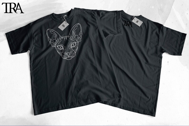 May include: Two black t-shirts with white graphic designs. One shirt features a line drawing of a Sphynx cat's head. The other shirt has the word "TRA" printed on the front.
