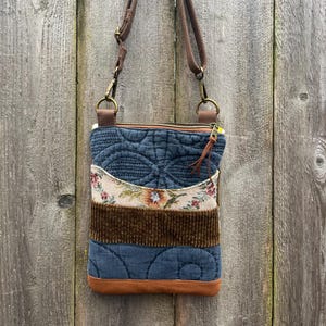 May include: A quilted crossbody bag with a brown strap and zipper. The bag features a patchwork design with blue denim, floral fabric, and brown corduroy. The bottom is trimmed with brown leather. The bag is designed for everyday use.