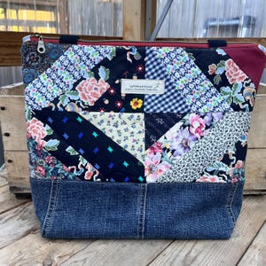 May include: A tote bag featuring a patchwork design of various floral, gingham, and solid fabric patterns in black, blue, and pink. The bag has a dark blue denim base, a top zipper, and a white label reading PATTERN & STITCH CO.