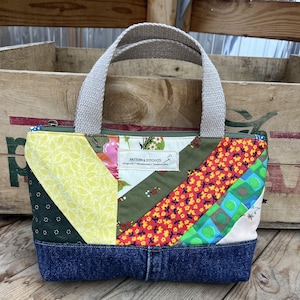 May include: A quilted fabric handbag featuring a patchwork design of yellow, green, red, and blue floral patterns. The bag has a dark blue denim base, beige woven handles, and a central white label reading PATTERN & STITCH CO.