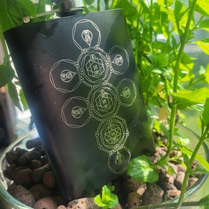 May include: Black metal flask with a silver cap and a geometric design etched on the front. The design features a series of circles and lines, with a central circle containing the letter "M".