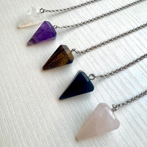 May include: Five crystal pendulums with silver chains. The crystals are clear, purple, brown, dark blue, and pink. Each pendulum is a triangular shape. The pendulums are arranged diagonally against a white background.