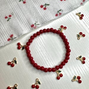 Op de afbeelding: Een rode kralenarmband met kersenbedels. De armband heeft kleine, ronde rode kralen en goudkleurige kersenbedels met rode kersen en groene bladeren. De achtergrond omvat een witte stof met een kersenpatroon en extra kersenbedels.