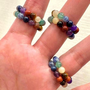 May include: Three stacked bracelets made of colorful gemstone beads. The bracelets are made of blue, green, red, brown, and purple beads.