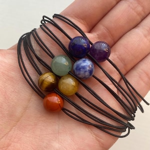 May include: A set of seven chakra bracelets with black elastic cord and round gemstone beads in red, orange, yellow, green, blue, indigo, and violet.