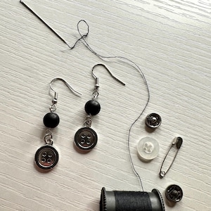 May include: A pair of handmade earrings with black beads and button charms. The image also shows sewing notions: a needle, thread, snaps, a safety pin, and a spool of black thread. The items are arranged on a white surface.