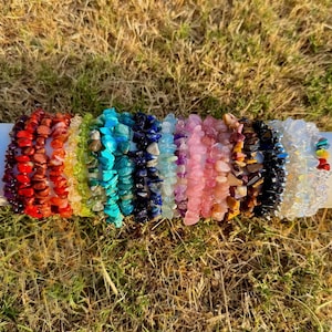 May include: A collection of 14 colorful gemstone bracelets, each made with small, irregular shaped stones. The bracelets are arranged in a rainbow order, starting with red and ending with white.