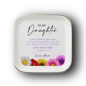 May include: A square white ceramic trinket dish with a gold-toned rim. The center features the text TO MY Daughter, followed by the Bible verse 1 Corinthians 13:4-8, and Love, Mom. A row of colorful daisy-like flowers lines the bottom edge.