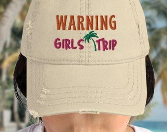 May include: A beige distressed baseball cap with the text "WARNING GIRLS TRIP" embroidered in orange, pink, and green. The text includes a palm tree graphic.