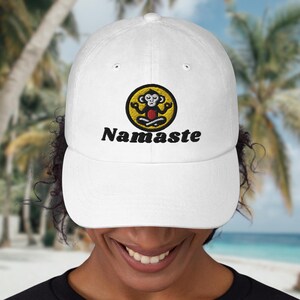 Può includere: Cappellino da baseball bianco con un cerchio giallo e una scimmia dei cartoni animati in una posa yoga. Il testo "Namaste" è ricamato sotto il cerchio.