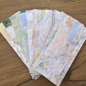 May include: A collection of folded paper maps, some with text like "Buffalo" and "Regina" visible. The maps are arranged in a fan shape on a wooden surface.