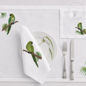 May include: White linen placemat, napkin, and coaster set with embroidered green parrots perched on branches. Includes a white plate with a floral design, silverware, and two wine glasses.