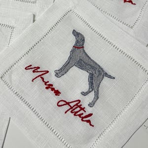 Puede incluir: Posavasos cuadrados de lino blanco con un diseño de perro gris bordado y el texto "Maison Attila" en rojo. Los posavasos tienen un borde cosido. Un conjunto de artículos de decoración.
