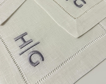 May include: A collection of off-white rectangular fabric napkins, each featuring a stitched border and the embroidered grey monogram H/G centred in the middle. The napkins are arranged in an overlapping pattern, showcasing the fabric texture.
