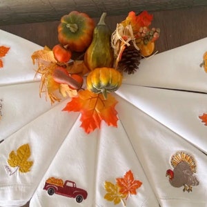 Op de afbeelding: Set van acht witte linnen servetten met geborduurde herfstthema-ontwerpen. De ontwerpen omvatten pompoenen, bladeren, een kalkoen en een vrachtwagen met pompoenen. De servetten zijn gerangschikt in een cirkel rond een middelpunt van herfstbladeren.