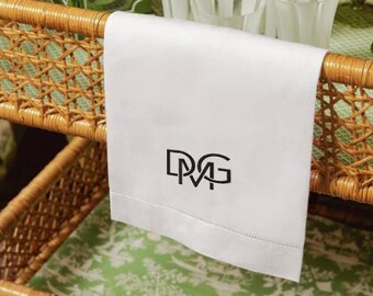 May include: A white hand towel with black embroidered initials "D.M.G."  The towel is folded and draped over a wicker basket.