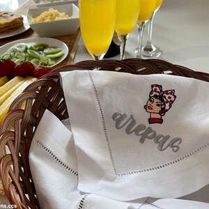 May include: White linen napkin with embroidered text "arepas" and a cartoon image of a woman with a red and white polka dot headband. The napkin is folded and placed in a wicker basket.