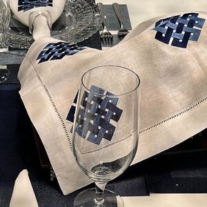 Könnte beinhalten: Eine weiße Leinen-Serviette mit einem blauen, gestickten Knotendesign. Die Serviette ist gefaltet und auf einem Tischgedeck mit einem Wasserglas und einem Platzdeckchen platziert.