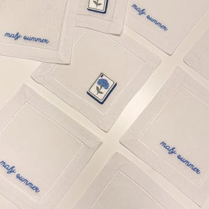 May include: A set of square white linen napkins featuring embroidered blue hydrangea flowers on rectangular patches and the blue cursive text "mafy summer" stitched along the edges, with hemstitched borders on each individual piece.