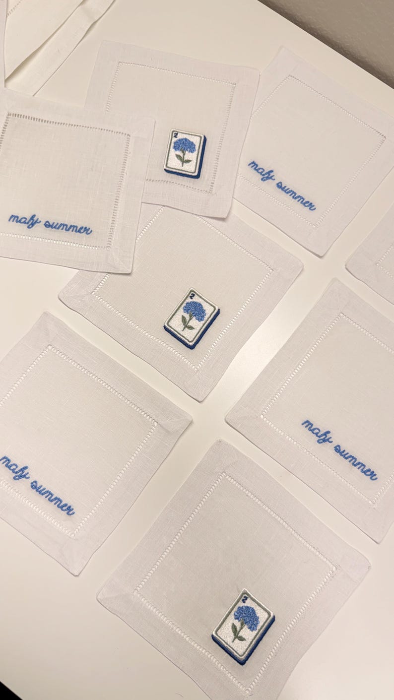 May include: A set of square white linen napkins featuring embroidered blue hydrangea flowers on rectangular patches and the blue cursive text "mafy summer" stitched along the edges, with hemstitched borders on each individual piece.