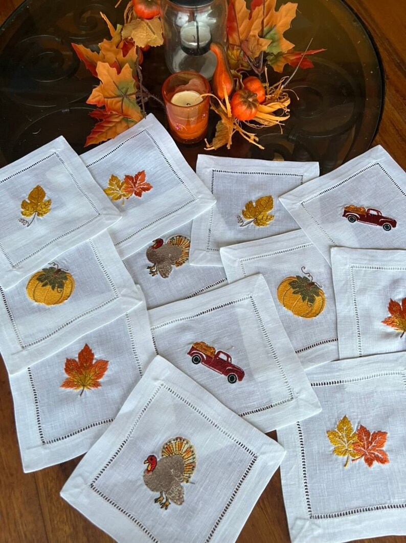 Op de afbeelding: Set van tien witte linnen servetten met geborduurde Thanksgiving-ontwerpen. De ontwerpen omvatten een pompoen, een kalkoen, een vrachtwagen met pompoenen en herfstbladeren in tinten oranje, geel en bruin.