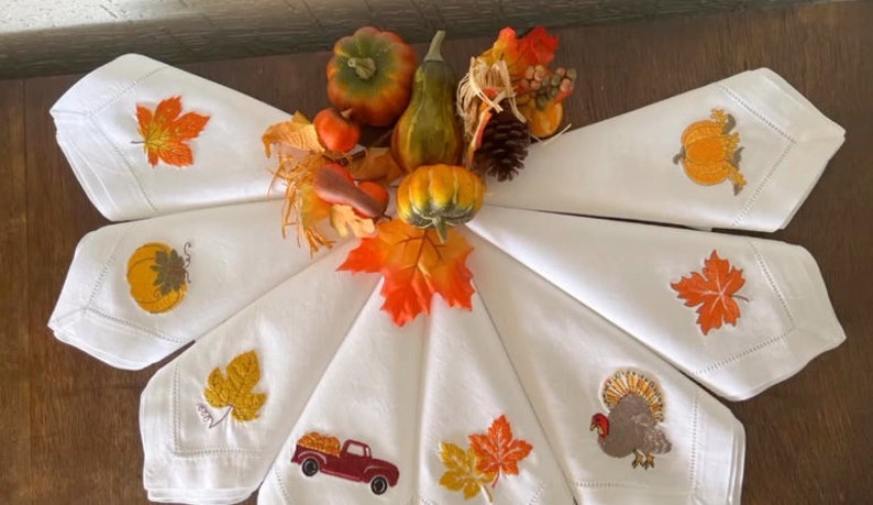 Op de afbeelding: Set van acht witte linnen servetten met geborduurde herfstthema-ontwerpen. De ontwerpen omvatten pompoenen, bladeren, een kalkoen en een vrachtwagen met pompoenen. De servetten zijn gerangschikt in een cirkel rond een middelpunt van herfstbladeren.