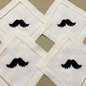 May include: Four white linen napkins with black embroidered moustaches. The napkins are arranged in a diamond pattern.